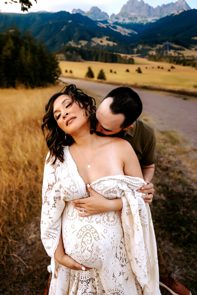 Man kissing on woman's neck while woman holds her baby bump during sunset