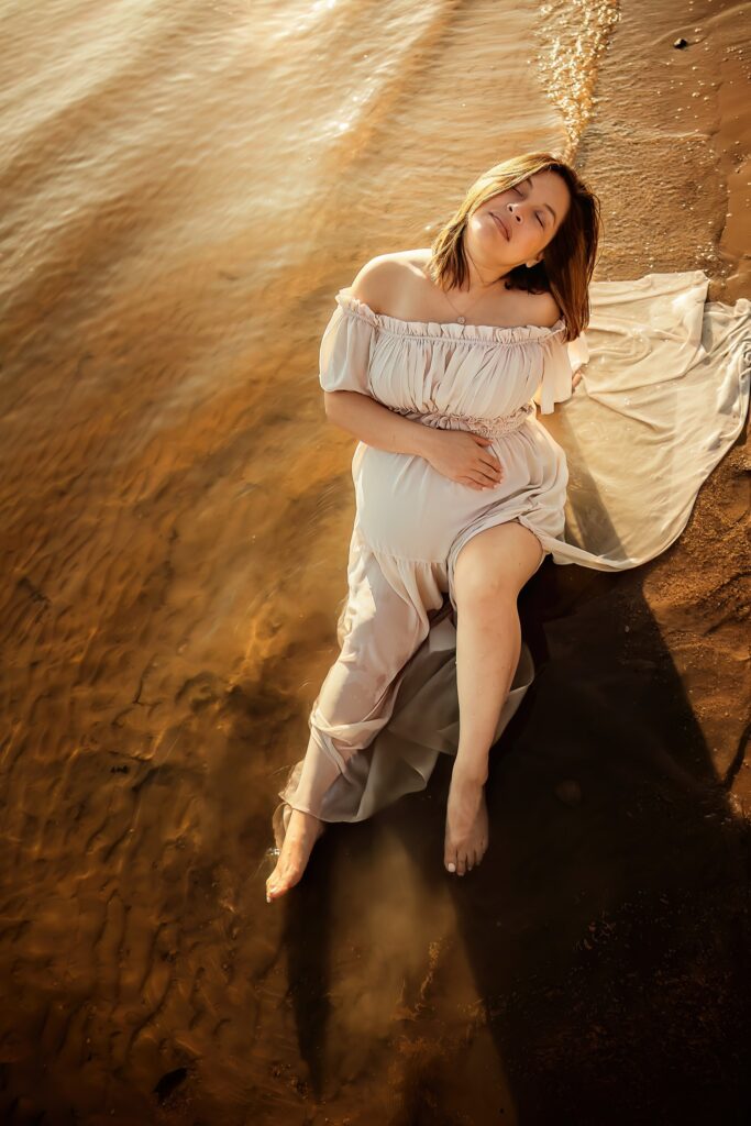 woman sits in a lake in a white dress after deciding what to wear for her maternity session.